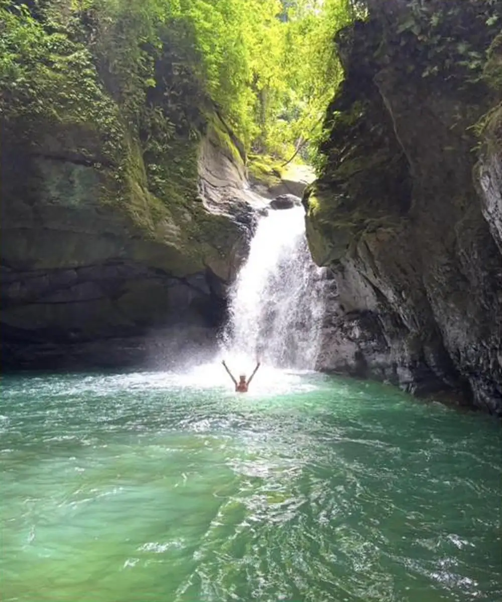 A guest standing beneath La Diosa waterfall with arms raised, turquoise pool below